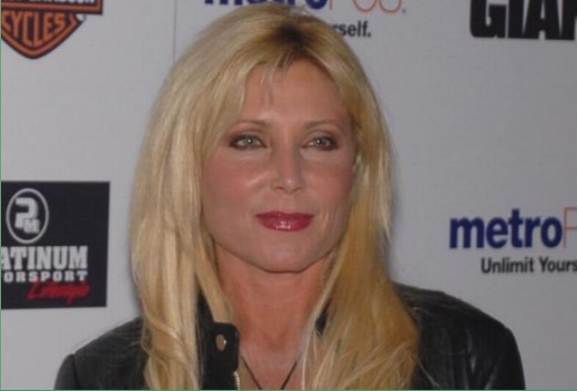 Pamela Bach posing at an event with a backdrop featuring various logos, including "metro" and "Platinum Motorsport.