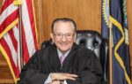 Elderly judge Frank Caprio wearing black robes and glasses, sitting at his bench with American flags visible in the background in a wood-paneled courtroom.