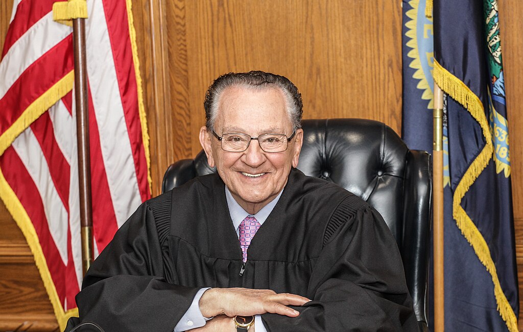 Elderly judge Frank Caprio wearing black robes and glasses, sitting at his bench with American flags visible in the background in a wood-paneled courtroom.
