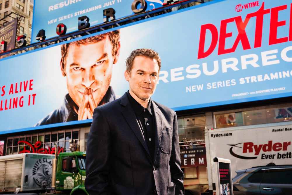 Michael C. Hall standing in front of a large Dexter: Resurrection billboard in Times Square, wearing a dark suit with the show's premiere date visible.