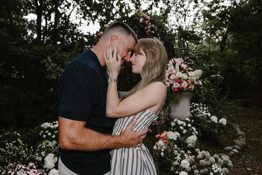 Taylor Swift and Travis Kelce embracing in a garden setting with flowers in the background, where he proposed. Swift wears a striped dress while Kelce is in a dark t-shirt as they touch foreheads intimately.