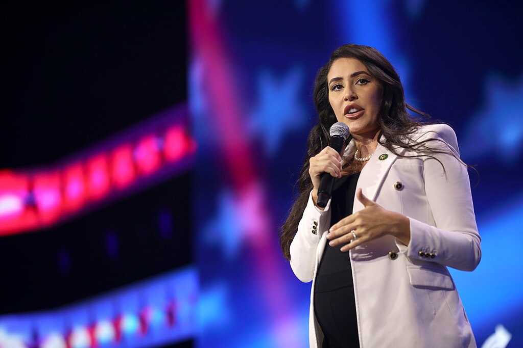 Florida Republican Representative Anna Paulina Luna speaking at a podium with a microphone, wearing a white blazer against a blurred blue and red background at the 2023 Turning Point Action Conference.