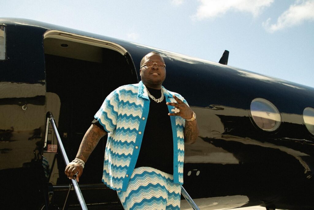 Sean Kingston wearing a blue and white zigzag pattern shirt standing on the steps of a private jet with jewelry and rings visible.