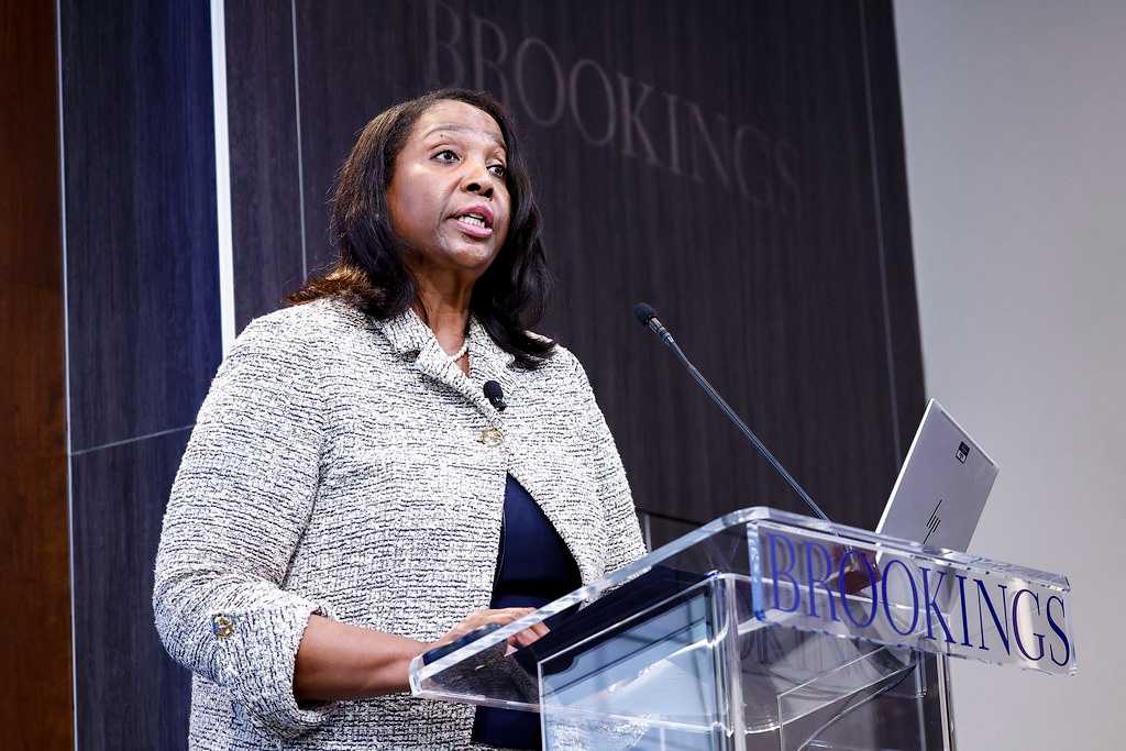 Federal Reserve Governor Lisa Cook giving a speech at a podium with the Brookings Institution logo visible in the background. She is wearing a white tweed jacket over a dark top and speaking into a microphone.