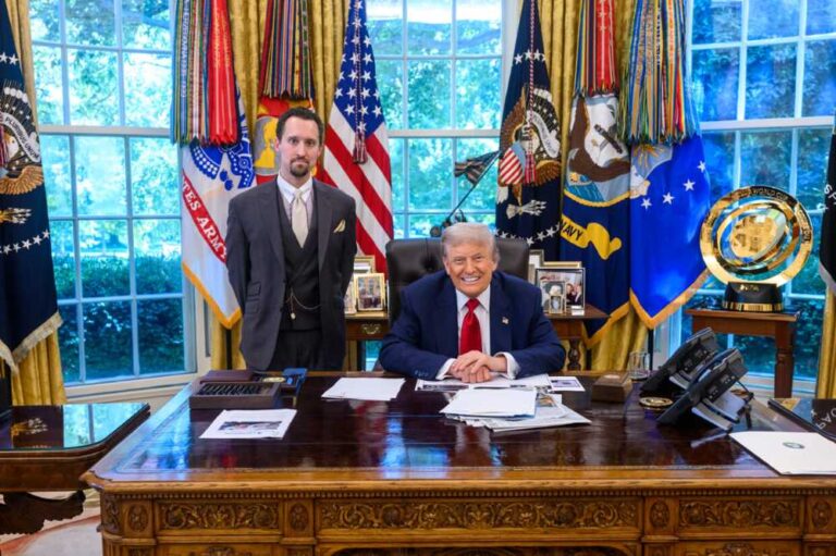 President Donald Trump sits at his desk in the Oval Office with E.J. Antoni standing next to him. Trump is wearing a dark blue suit with a red tie, while Antoni is dressed in a dark suit with a yellow pocket square. American flags and military flags are visible in the background.
