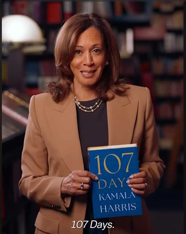 Kamala Harris stands in front of a bookshelf, smiling and holding up a blue hardcover titled “107 Days” as she addresses the camera.
