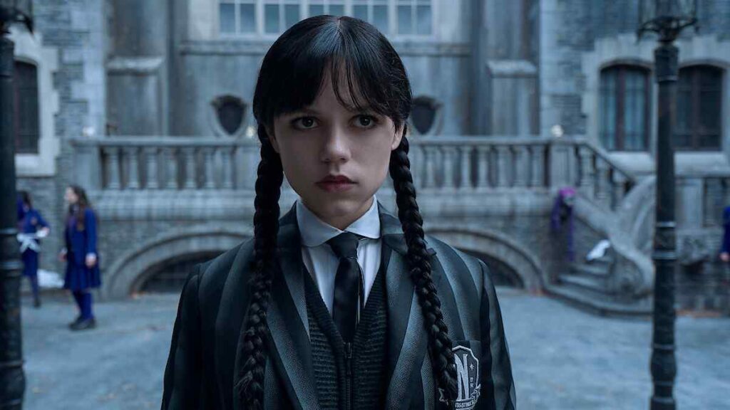 Jenna Ortega as Wednesday Addams, dressed in her Wednesday-themed school uniform with braided pigtails, standing before the gothic stone façade of Nevermore Academy.