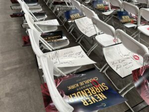 White folding chairs at State Farm Stadium set up for Charlie Kirk's memorial service, with handwritten notes quoting Isaiah 6:8 and "Never Surrender" promotional materials placed on seats before the event begins.