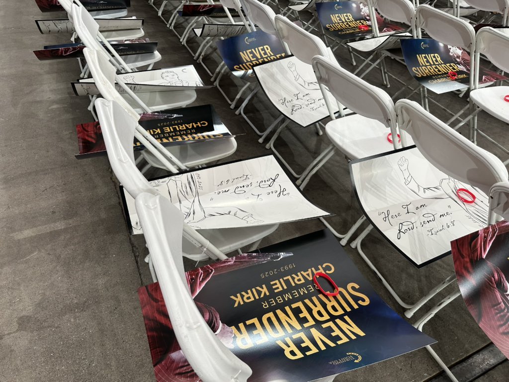 White folding chairs at State Farm Stadium set up for Charlie Kirk's memorial service, with handwritten notes quoting Isaiah 6:8 and "Never Surrender" promotional materials placed on seats before the event begins.