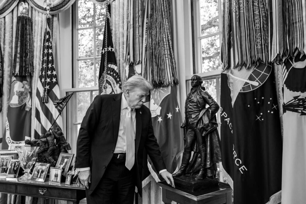 Representative Image. Black and white photograph of former President Donald Trump standing in the Oval Office beside a statue, with American flags and decorative curtains visible in the background.