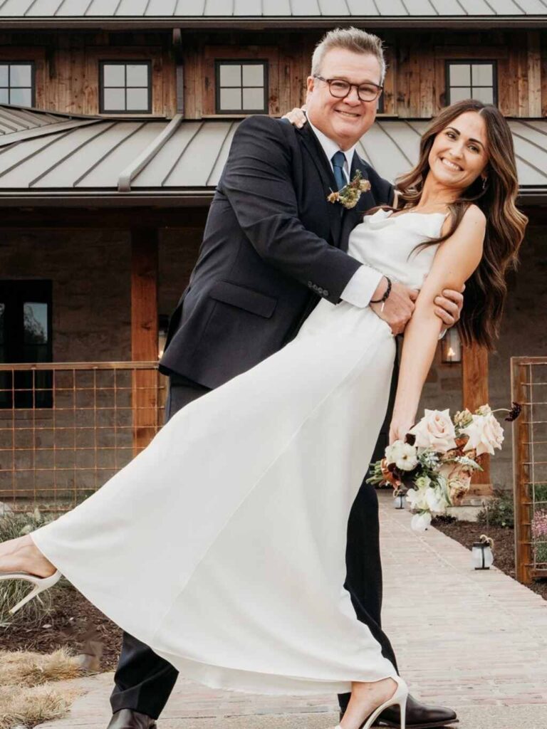 Eric Stonestreet wearing a navy suit holds Lindsay Schweitzer in a white wedding gown outside their wooden home with a metal roof, both smiling at the camera.