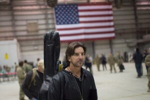 Brett James walking with a guitar case slung over his shoulder, standing in front of an American flag backdrop.