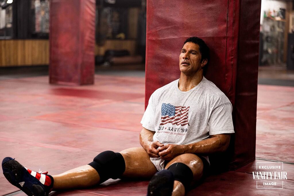 Dwayne Johnson as Mark Kerr sitting against a red punching bag in a gym, wearing a white t-shirt with an American flag and "U.S. Navy '90" printed on it, looking exhausted and contemplative after training.