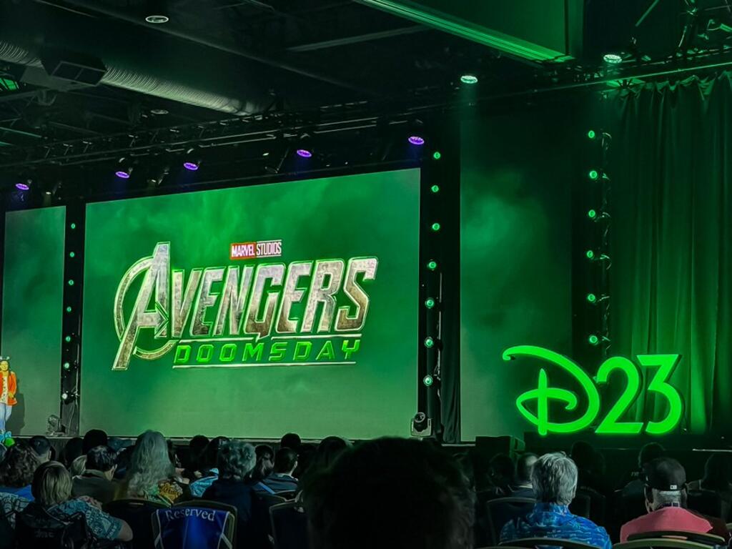 The Avengers: Doomsday logo displayed on a large screen with green lighting at Disney's D23 event, showing the Marvel Studios branding above the title and a D23 logo visible on the right side of the stage, with audience members visible in the foreground.