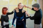 Sophie Turner getting her blonde hair styled by two stylists wearing black, one applying product while she smiles in a dark blue velvet dress.