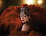 Taylor Swift in glamorous showgirl-inspired styling featuring a diamond headpiece and red feathered costume against a dark background with spotlights, promoting her new album "The Life of a Showgirl"