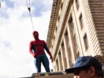 Tom Holland in Spider-Man costume suspended on wires during filming of Spider-Man Brand New Day with crew member visible below on building exterior