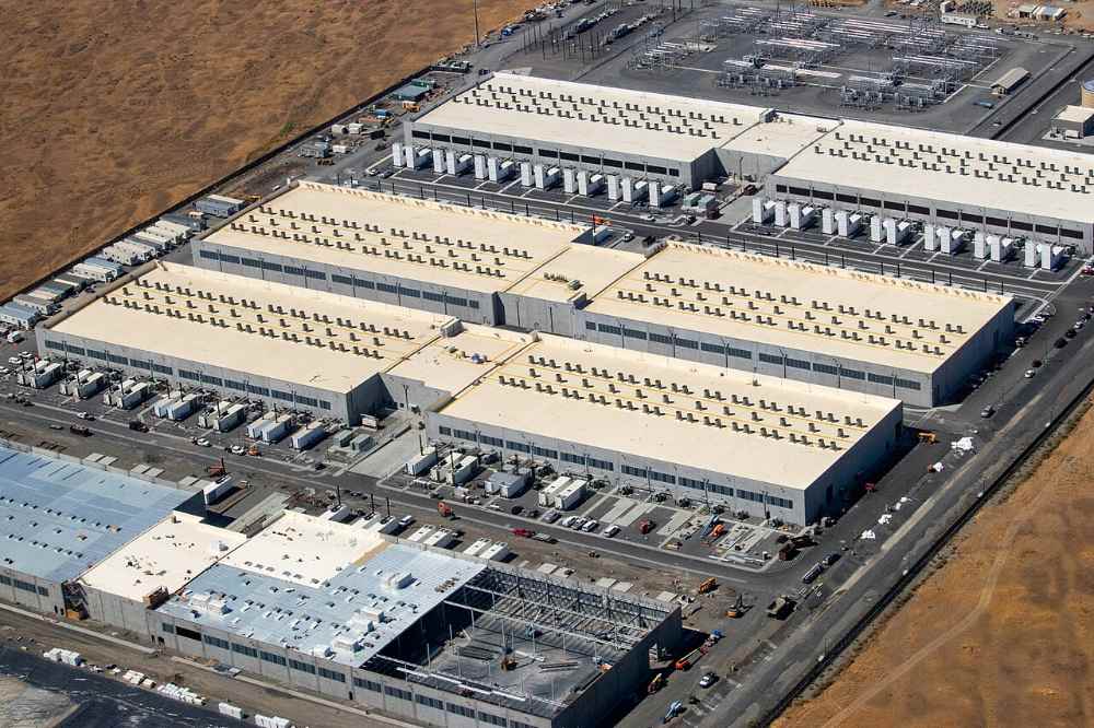 Wide view of an Amazon Web Services data center site in the us-west-2 region, Morrow County, Oregon, showing three large warehouse-style buildings, a fourth building under construction with cranes, electrical substations, and transmission lines in a semi-arid landscape.