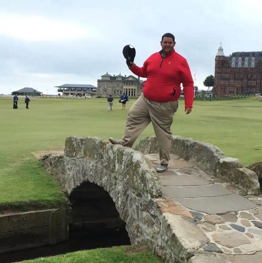 Cody “Beef” Franke stands on the Swilcan Bridge at the Old Course, St Andrews, holding his cap in his right hand and wearing a red quarter-zip and khaki pants; the R&A clubhouse and surrounding buildings are visible in the background with players on the fairway.