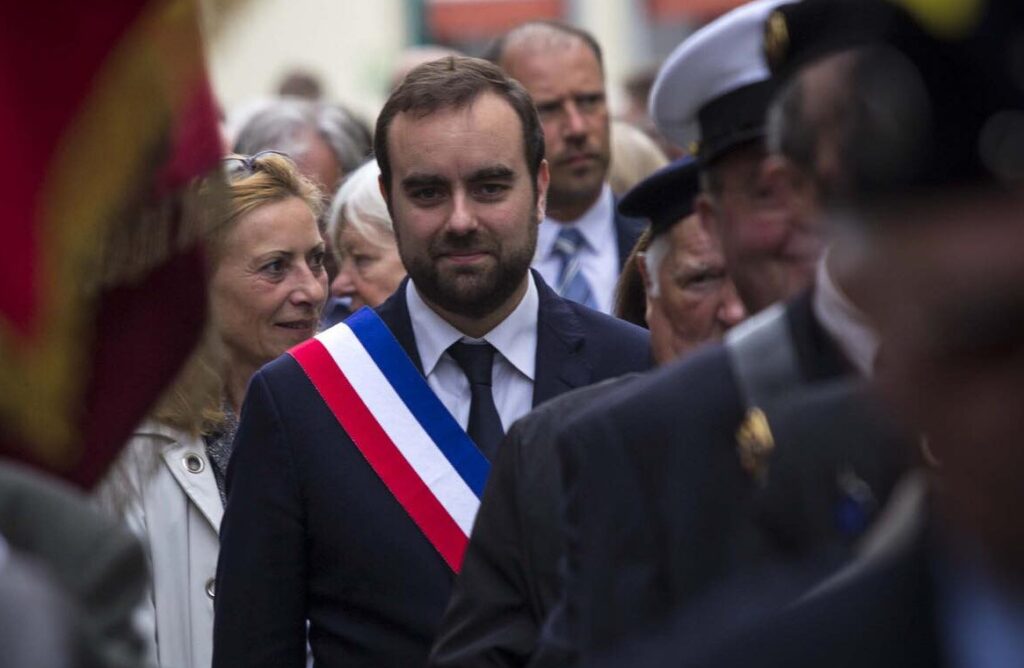 A man in a suit with a blue, white, and red sash, smiling slightly, surrounded by other people in a blurry outdoor setting, possibly during a parade or public event.