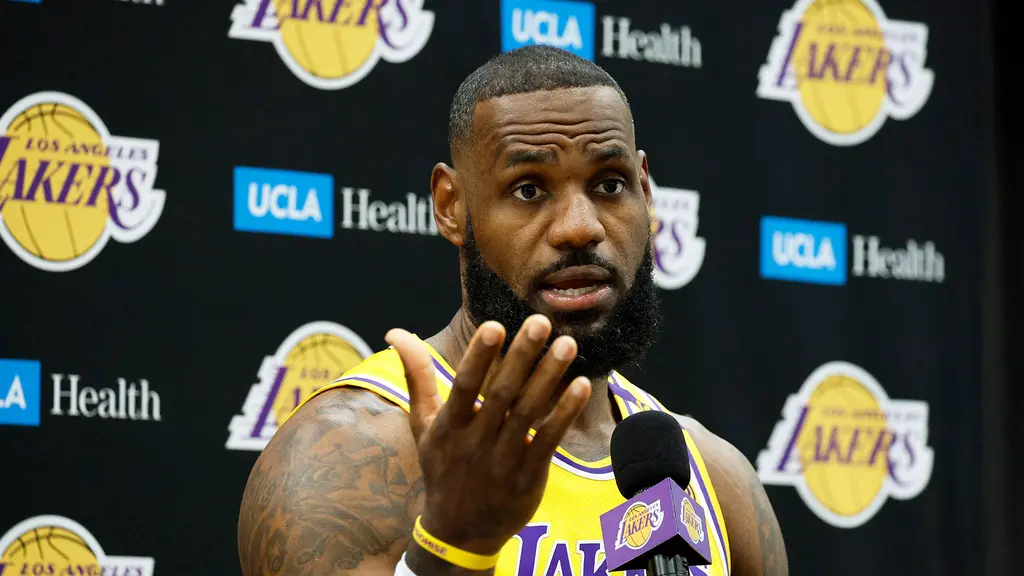 LeBron James speaks into a microphone at a press conference with Lakers and UCLA Health logos in the background.