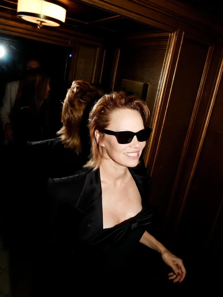 Pamela Anderson walking through a mirrored hallway in Paris, wearing black sunglasses and showcasing her voluminous copper hairstyle