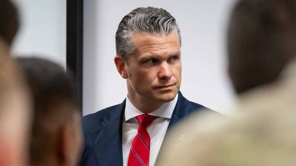 Pete Hegseth in a dark suit and red tie during a briefing, with blurred uniformed service members in the foreground.
