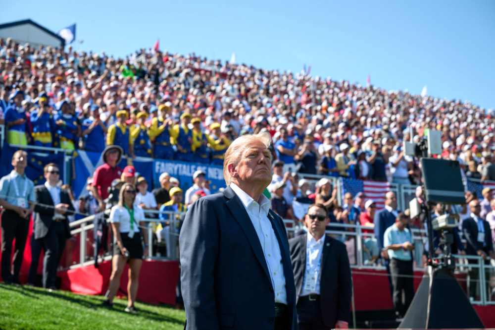 Donald Trump stands at an outdoor event in front of a large crowd with U.S. flags visible, midday light, security staff nearby.