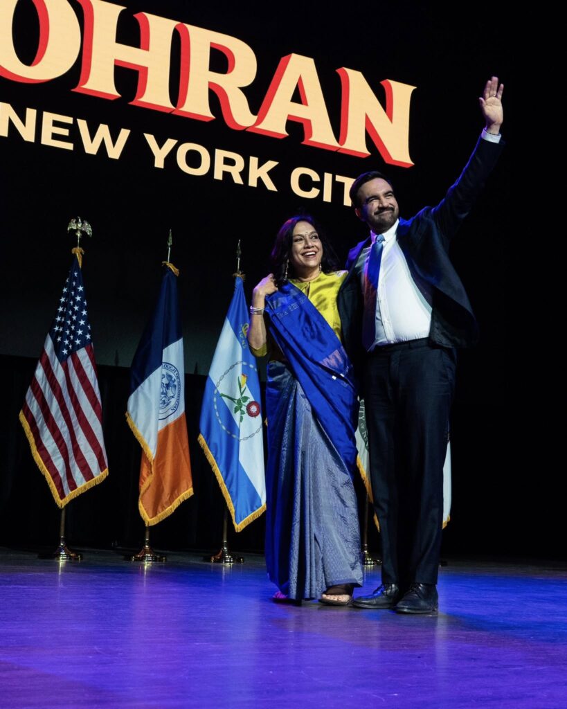 Stage photo at NYC election-night event showing Mira Nair in a blue saree and Mamdani waving, with U.S., New York City, and campaign flags behind them.