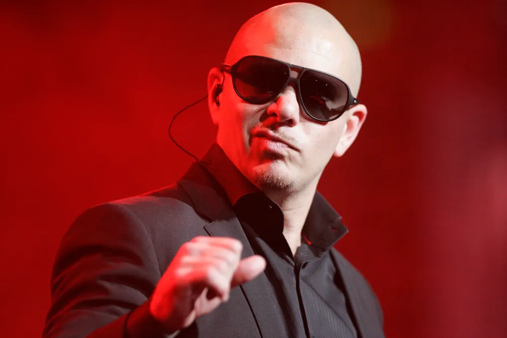 Pitbull performing live on stage in 2012 wearing a black tuxedo suit and sunglasses, holding a microphone with a determined expression, serving as the visual inspiration for the 2026 bald cap fan trend.
