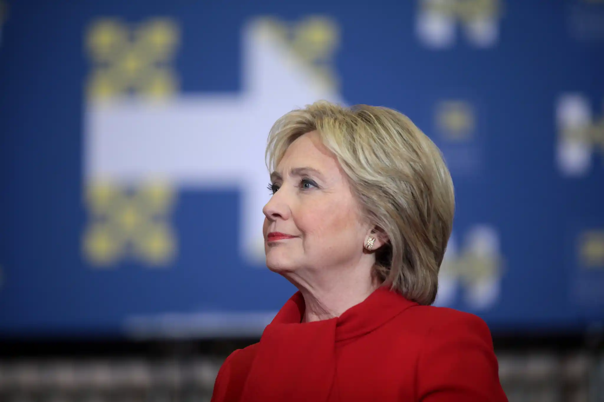 Hillary Clinton in a red outfit speaking at a campaign event with a blue backdrop featuring an “H” logo in Iowa.
