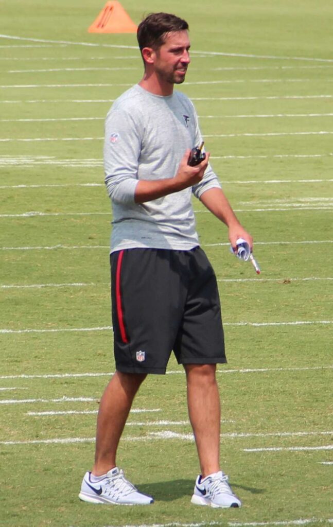 Kyle Shanahan wearing a red Atlanta Falcons polo shirt and sunglasses, speaking during Falcons training camp in July 2016.