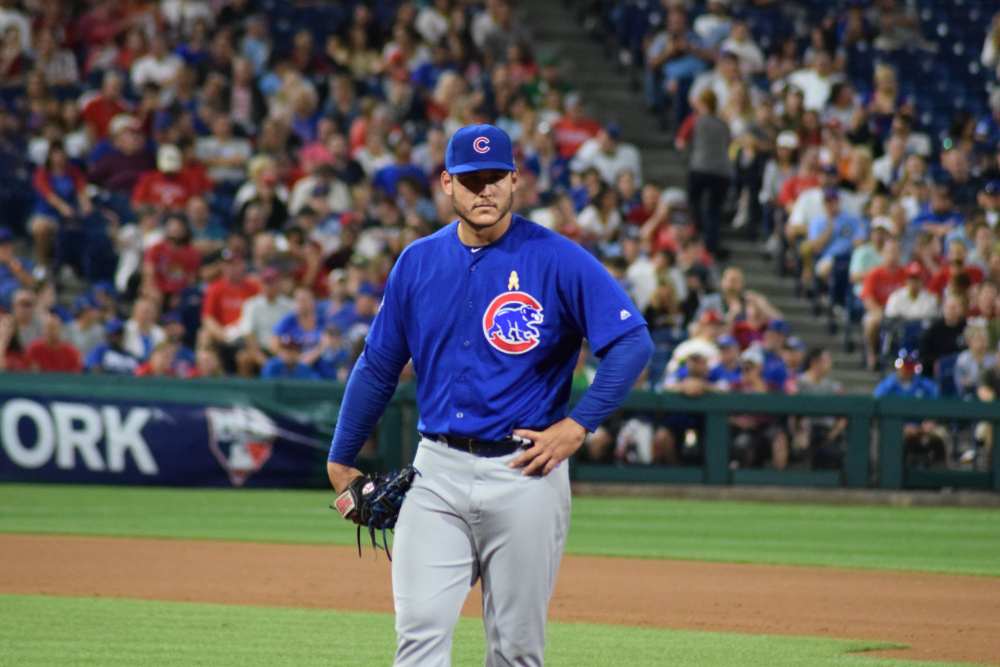 Anthony Rizzo, wearing a navy blue button-down shirt, stands in the Wrigley Field bleachers with his right arm raised, holding a baseball, while a young boy in a Cubs jersey sits on his shoulders against a backdrop of cheering fans.