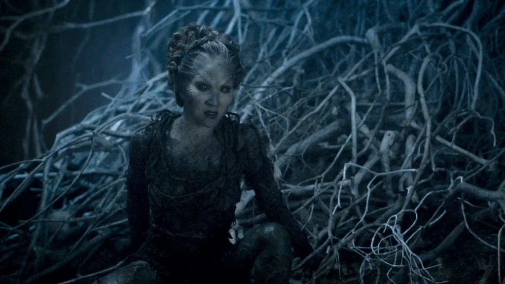 Supernatural character with pale complexion and dark eyes surrounded by frost-covered branches and mystical forest vegetation in a Game of Thrones scene.