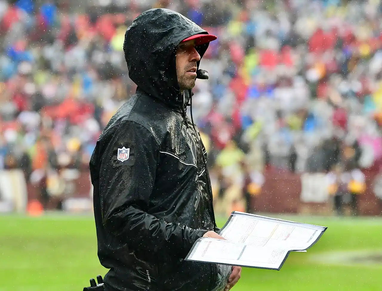 Kyle Shanahan, San Francisco 49ers head coach, standing on sideline during rainy game conditions holding laminated playbook with headset communication device.